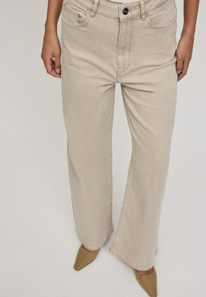 Personne portant un jean beige clair à jambes larges et des chaussures à bout pointu couleur fauve, debout avec les mains détendues le long du corps sur un fond neutre.