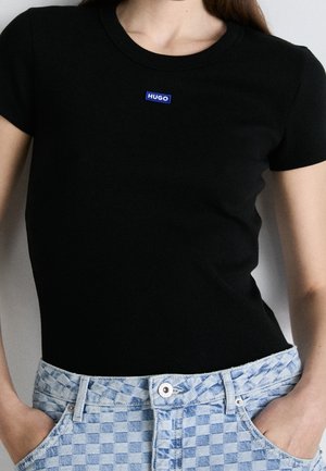 Person iført en sort kortærmet skjorte med et lille blåt "HUGO"-logo på brystet og lyseblå ternede jeans med hænderne i lommerne.