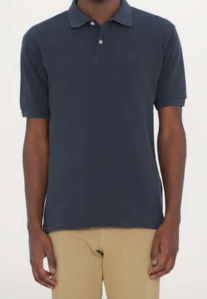 Homme portant un polo bleu marine foncé à manches courtes avec deux boutons et un pantalon beige, debout sur un fond clair uni.
