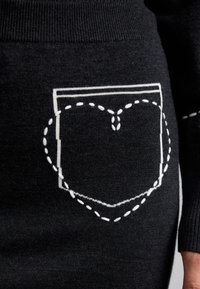 Îmbrăcăminte tricotată neagră, având un buzunar decorativ cu margine albă și un model de cusătură în formă de inimă. Textură fină cu manșete ribbate.