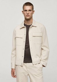 Light beige jacket with textured fabric, featuring a collared design, two front pockets, and silver snap buttons. Paired with a dark top.