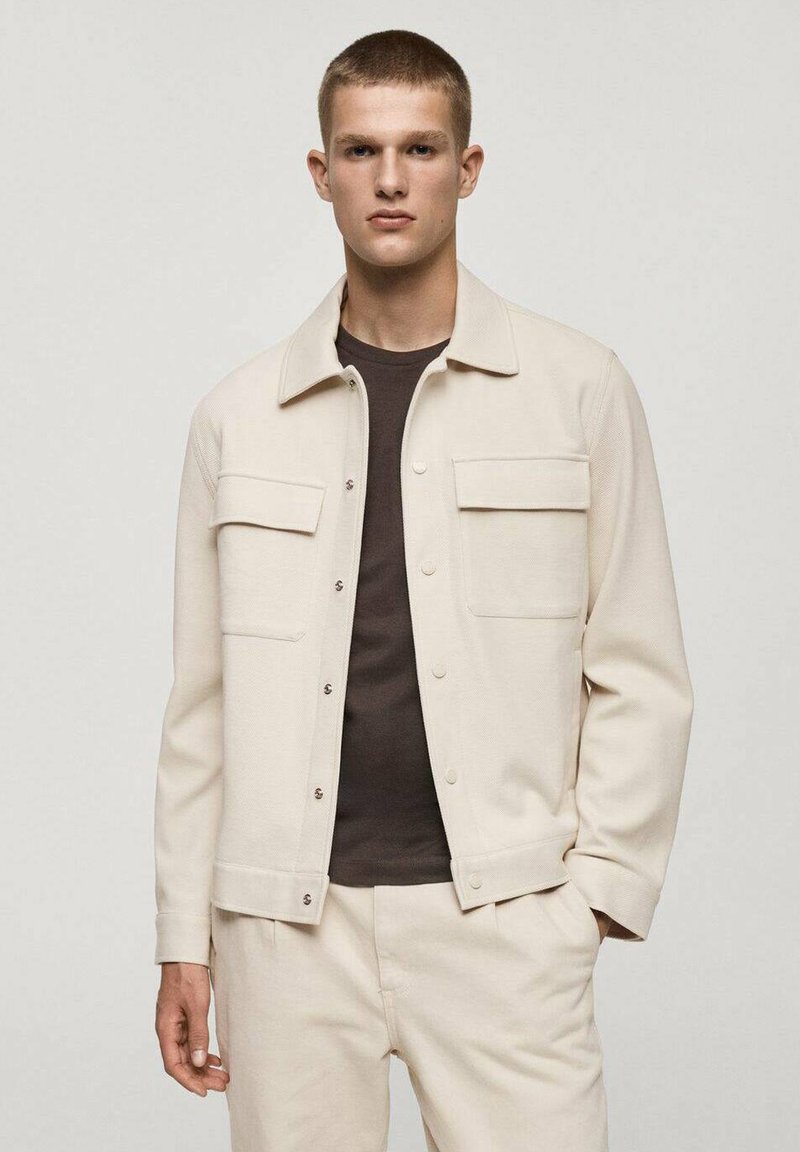 Light beige jacket with textured fabric, featuring a collared design, two front pockets, and silver snap buttons. Paired with a dark top.