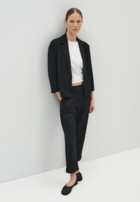 Blazer noir ajusté avec revers en pointe, t-shirt blanc court et pantalon noir. Le modèle porte des ballerines noires et pose sur un fond clair.