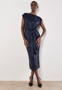 Granatowa sukienka z cekinami, z dekoltem w łódkę i krótkimi rękawami, związana w talii. Modelka zestawiła ją z czarnymi szpilkami w punkcie.