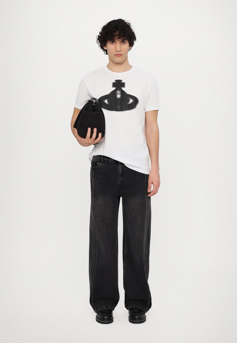 Weißes T-Shirt mit schwarzem Grafikdesign, kombiniert mit lockeren schwarzen Denim-Hosen und klobigen schwarzen Schuhen, während eine schwarze Tasche gehalten wird.