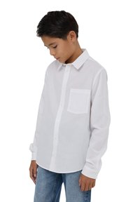 Ragazzo con capelli scuri che indossa una camicia bianca a maniche lunghe e jeans blu, guardando in basso su uno sfondo bianco.
