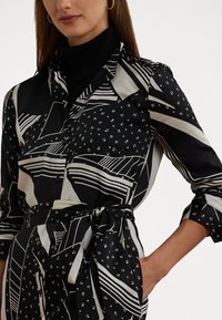 Svartvitt mönstrad skjortklänning med geometriska mönster, midjebälte, långa ärmar och en slät tygtextur.