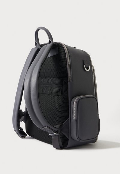 Sac à dos noir en nylon avec bretelles rembourrées, panneau arrière en mesh, poche avant zippée et détails de fermeture éclair argentés sur fond blanc.