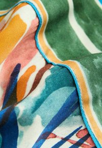Tissu coloré avec des motifs floraux abstraits en vert, orange et bleu ; présente un bord cousu bleu, une texture douce et un drapé fluide.