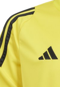 Geel sporttop gemaakt van gladde stof. Heeft zwarte strepen op de schouders en een zwarte geborduurde logo op de borst.