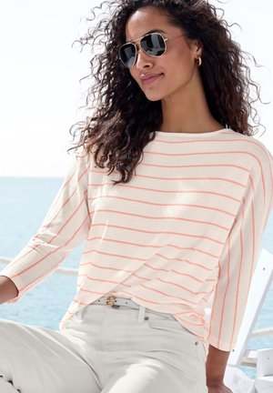 Leichtes, gestricktes Shirt mit weißen und korallenfarbenen horizontalen Streifen; verfügt über einen runden Halsausschnitt und drei Viertelärmel. Getragen mit weißen Hosen und einem Gürtel.