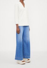 Vit långärmad toppen med V-hals och en enda knapp; kombinerad med vida, urblekta blå jeans och öppna vita klackskor.