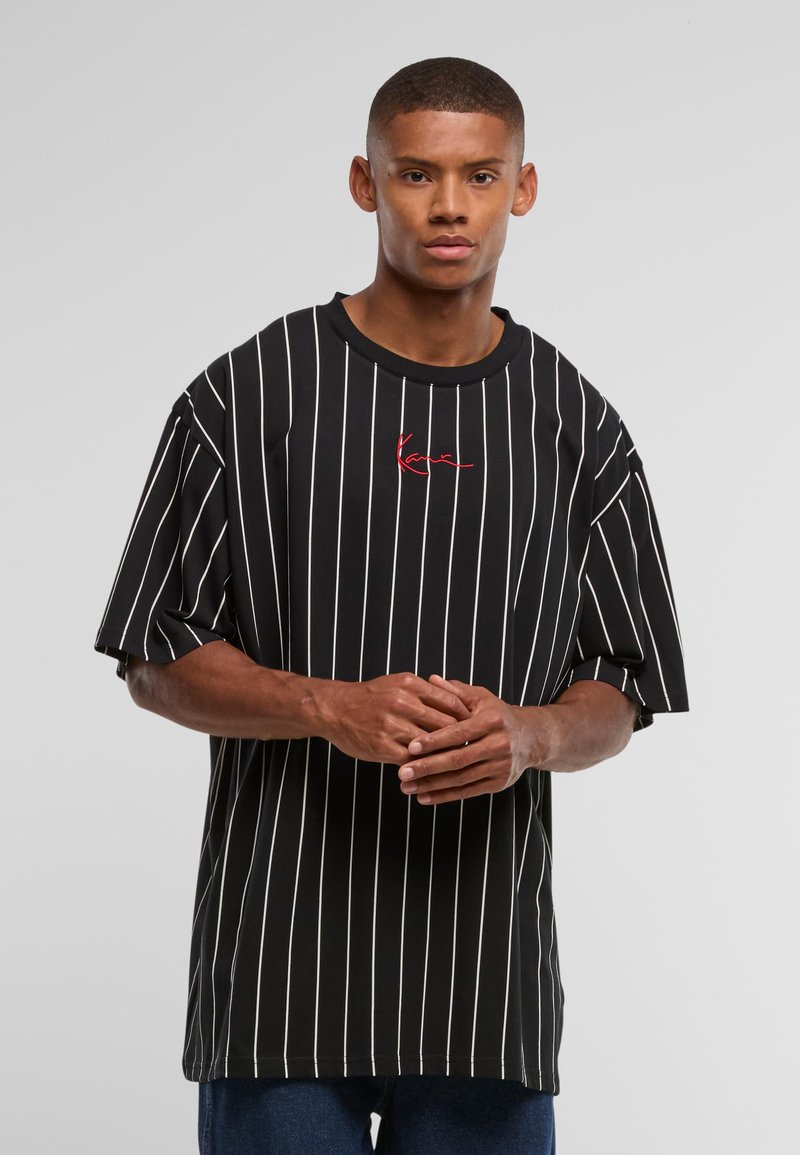 Junger Mann, der ein schwarzes, übergroßes T-Shirt mit weißen vertikalen Streifen und rotem Text trägt, vor einem schlichten hellgrauen Hintergrund steht.