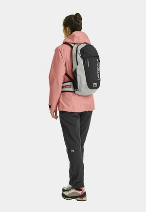 Gray and black backpack with geometric patterns, worn over a pink jacket and black pants, featuring adjustable straps and logo detail.