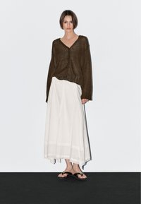 Bruine, oversized gebreide cardigan met een V-hals en knopen. Witte, luchtige maxi-rok met een getextureerde zoom. Zwarte teenslippers. Minimalistische achtergrond.