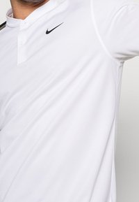 Bílá polokošile s límečkem na knoflíky, s černým logem Nike na levé straně hrudi, vyrobená z hladké, prodyšné látky.