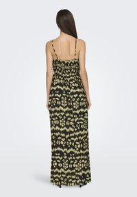 Mujer con cabello largo y lacio de color castaño, vistiendo un vestido maxi sin mangas de patrón negro y amarillo, de pie de espaldas.