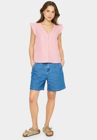 Blusa senza maniche a righe rosse e bianche con scollatura a V e spalline arricciate, abbinata a pantaloni shorts in denim blu e sandali beige.