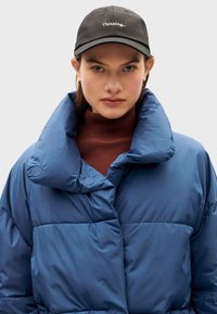 Chaqueta de plumas azul con un alto cuello y grandes secciones acolchadas. Lleva puesta sobre un cuello vuelto marrón. Gorro negro con el texto "Thinking...".