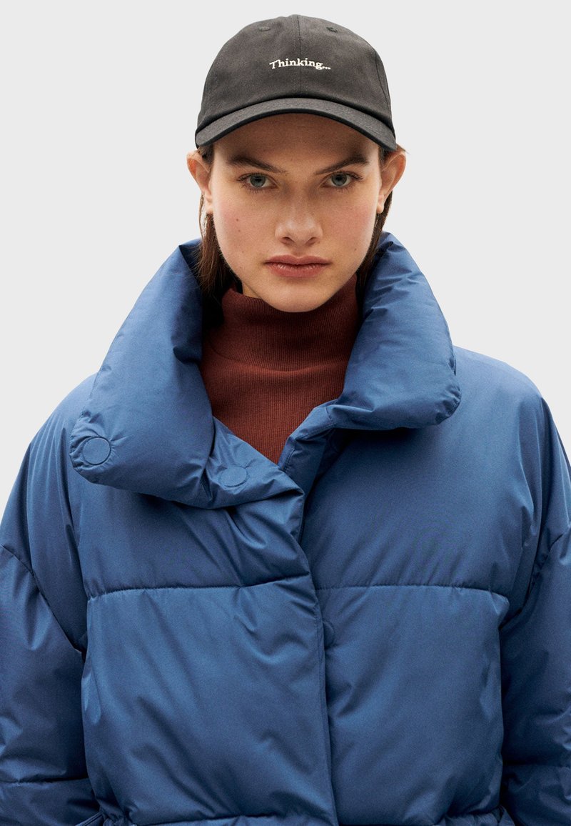 Chaqueta de plumas azul con un alto cuello y grandes secciones acolchadas. Lleva puesta sobre un cuello vuelto marrón. Gorro negro con el texto "Thinking...".