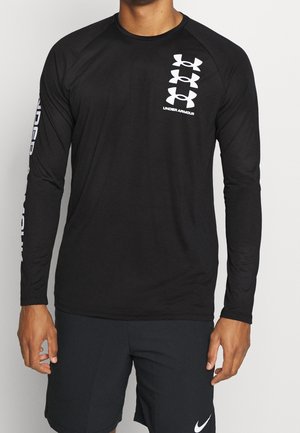 Mężczyzna w czarnej koszulce z długim rękawem Under Armour z białymi logo na klatce piersiowej i rękawie oraz czarnych spodenkach Nike z białym logo na lewej nodze.