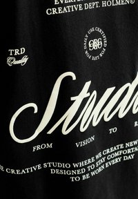 Tissu noir avec texte blanc comprenant « Studio », « Creative Dept. Holmen » et des phrases sur le design, le confort et la certification pour un usage quotidien.