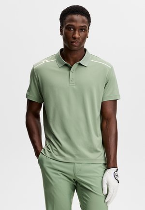 Mężczyzna ubrany w jasnozieloną koszulkę polo z krótkim rękawem i spodnie, stojący z jedną ręką w kieszeni i białą rękawiczką golfową na drugiej ręce.