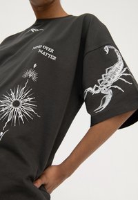 Person trägt ein schwarzes T-Shirt mit weißem Schriftzug "Mind Over Matter", Sonnenstrahl-Design auf der Vorderseite und einer detaillierten weißen Skorpion-Grafik auf dem Ärmel.