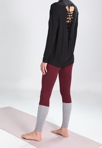 Top nero a maniche lunghe con dettaglio di lacci sul retro, abbinato a leggings color block bordeaux e grigi. Modella scalza su un tappetino.