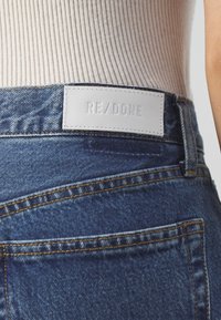 Vue arrière d'une personne portant un jean en denim bleu avec une étiquette blanche rectangulaire de la marque RE/DONE près de la taille, et un haut blanc côtelé.