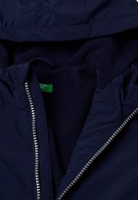 Námořnická modrá bunda s měkkou fleecovou podšívkou, předním zipem a vnitřním stahováním. Hladký vnější materiál a kontrastní vnitřní textura.