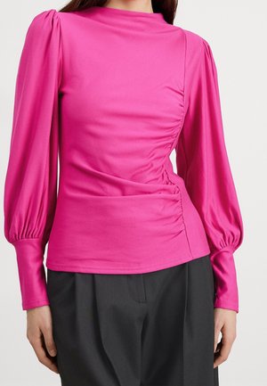Maglietta lunga rosa con colletto alto, caratterizzata da dettagli arricciati su un lato e una texture liscia. Indossata con pantaloni neri.