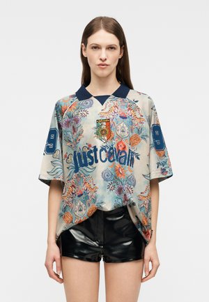 Γυναίκα που φορά μια πολύχρωμη, φαρδιά πουκάμισο με λουλουδάτο και τίγρη μοτίβο, με το κείμενο "just cavalli" και μπλε σκούρο γιακά, συνδυασμένο με μαύρο γυαλιστερό σορτς.