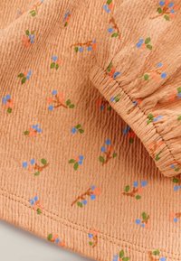 Tissu texturé de couleur pêche avec de petits motifs floraux en rose, bleu et vert. Présente un élastique froncé à l'ourlet.