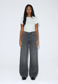 Frau mit langen schwarzen Haaren steht frontal, trägt ein weißes kurzärmeliges Shirt und hoch taillierte, weitgeschnittene graue Jeans mit schwarzen Schuhen.