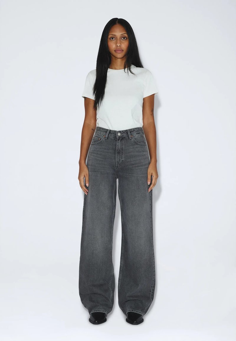 Woman with long black hair stands facing forward wearing a white short-sleeve shirt and high-waisted, wide-leg gray jeans with black shoes.