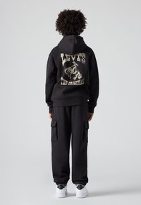 Černá mikina a kalhoty, oba vyrobené z měkkého materiálu. Mikina má na zádech zlaté logo a grafiku "Levi's". Bílé tenisky.