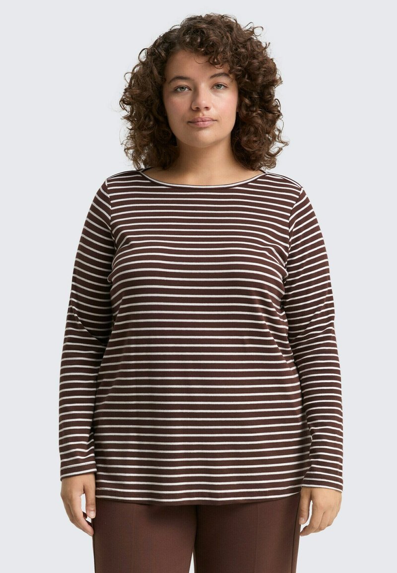 Frau mit lockigem Haar, die ein langärmliges braun-weiß gestreiftes Hemd und braune Hosen trägt, und gegen einen schlichten hellen Hintergrund steht.