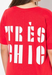 T-shirt rouge à coupe ample avec un grand texte blanc au dos qui dit "TRÈS CHIC". Texture de tissu douce avec un design décontracté.
