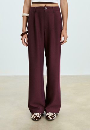 Donna che indossa un pantalone largo a vita alta color mirtillo, una maglietta bianca corta, delle scarpe a stampa mucca e braccialetti spessi su entrambi i polsi.
