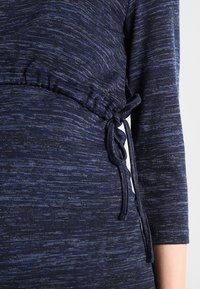 Mörkblå stickad plagg med en texturerad mönster, med en åtsittande midja med en knytdetalj och ärmar som når tre fjärdedelar.