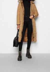 Lång, pösig brun jacka med huva och pälsdetaljer, tillsammans med åtsittande svarta jeans och svarta kängor med gyllene öglor och tjocka sulor.