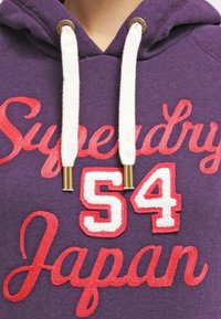Sudadera morada con bordado rojo. Presenta el texto "Superdry 54 Japan", cordones blancos con herrajes dorados en el cuello. Textura de tela suave.