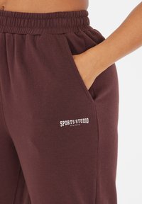 Burgunderfarbene Sweatpants mit elastischem Bund, Seitentaschen und dem Aufdruck "SPORTS STUDIO ATHLETICA" in Weiß auf dem linken Oberschenkel.