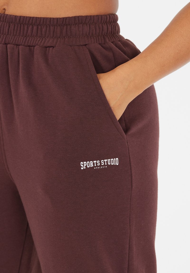 Burgunderfarbene Sweatpants mit elastischem Bund, Seitentaschen und dem Aufdruck "SPORTS STUDIO ATHLETICA" in Weiß auf dem linken Oberschenkel.