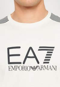 Balta medvilninė marškinėlių su dideliu juodu "EA7" logotipu ir "EMPORIO ARMANI" tekstu, papuošti juodomis juostelėmis ant pečių.