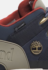 Granatowe i szare trampki z fakturowaną cholewką, czerwoną wyściółką, złotym logo drzewa oraz etykietą TimberDry, zapewniającą wodoodporność.