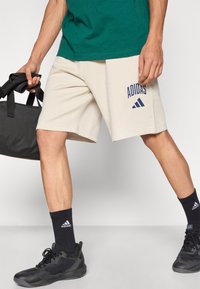 Μπεζ σορτς Adidas με πλαϊνές τσέπες και μπλε σκούρο λογότυπο, συνδυασμένα με μαύρες κάλτσες και παπούτσια Adidas. Το μοντέλο κρατάει μια μαύρη τσάντα.