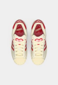 Κόκκινα και κρεμ αθλητικά παπούτσια με υφασμένη λαστιχένια μύτη, τρεις κόκκινες ρίγες και διακοσμητικά σε σχήμα καρδιάς. Το εσωτερικό διαθέτει σχέδιο πλέγματος.