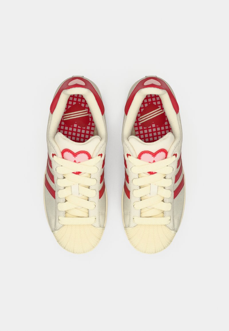 Baskets rouges et crème avec un embout en caoutchouc texturé, trois bandes rouges et des détails en forme de cœur. L'intérieur présente un motif en grille.